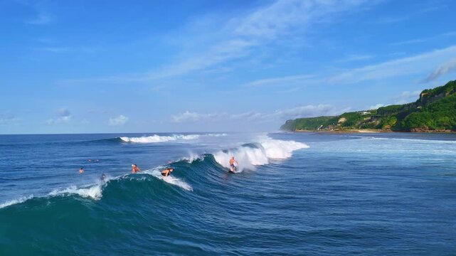 Aerial drone shot flying parallel to surfers riding waves near dramatic rocky cliffs in Bali, Indonesia. Dynamic action scene capturing the thrill of surfing in crystal clear ocean water