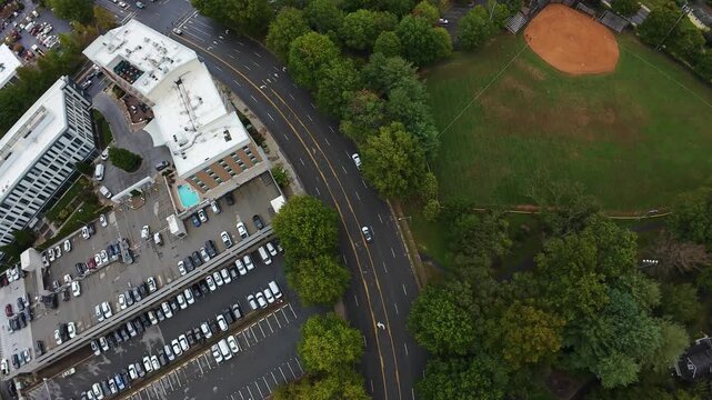 Aerial Asheville Ballfield Beside Commercial Parking Lot, Curved Road Frames Green Diamond Infield, Adjacent Buildings With Rooftop Pool, Tree Belt And Commuter Lanes, Community Recreation