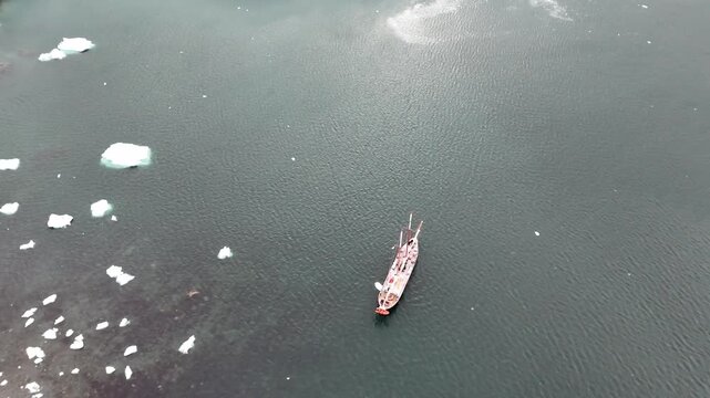 Cinematic descending over the icy waters of Melchior Harbour to reveal a top-down close-up of the wooden deck and rigging of the expedition schooner Amazone in Antarctica