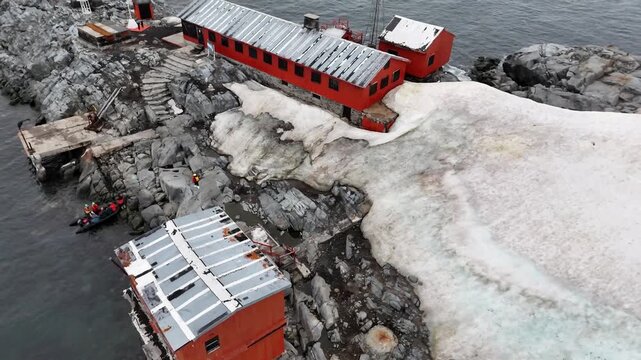Aerial raises over the historic red buildings of Argentina's Melchior Base to reveal tourists in a black Zodiac inflatable boat departing the rocky shores of Gamma Island, Antarctica