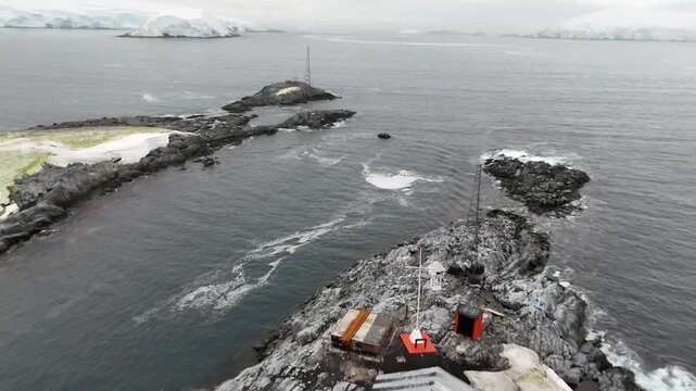 Cinematic aerial moving from the abandoned antenna towers of Argentina's Melchior Base to follow a black Zodiac boat as it carries expedition passengers across the dark waters of the Southern Ocean