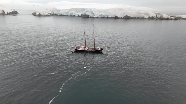 Cinematic aerial moving forward over the dark waters of Melchior Harbour toward the expedition schooner Amazone, framed by the blue ice cliffs and volcanic rocks of Gamma Island, Antarctica
