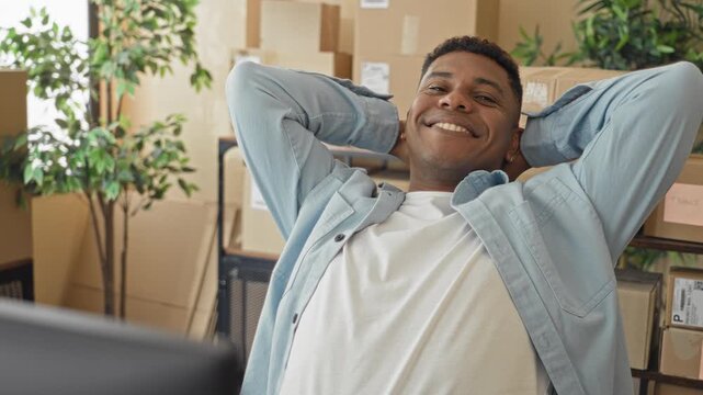 Man leaning back with hands behind head holding a parcel by a computer desk in a building packed with cardboard boxes; relief success.
