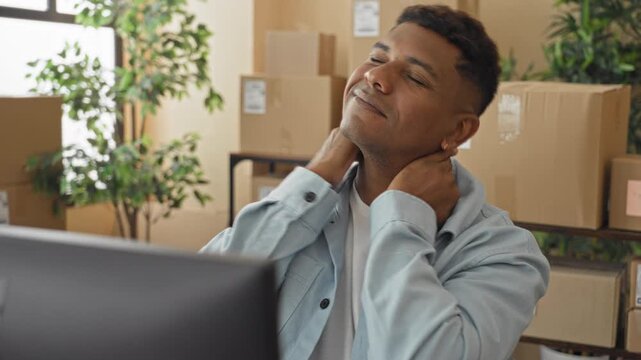 Man rubbing neck and shoulders by computer amid stacked shipping boxes in light blue shirt in building; fatigue relief.