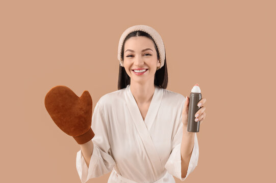 Happy young woman in bathrobe with massage glove and bottle of self tanner mousse on beige background