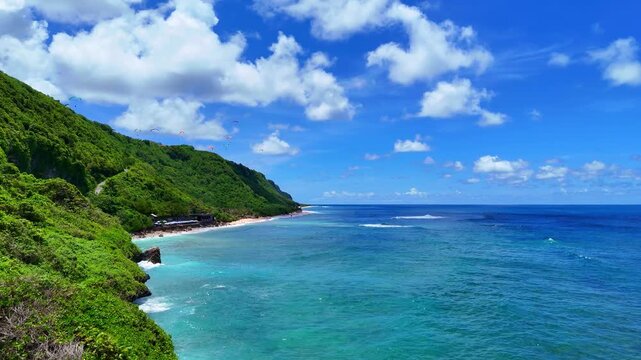 Breathtaking drone footage soaring over turquoise ocean waters along dramatic limestone cliffs in Uluwatu, Bali. Lush green hills and rugged coastline with crashing waves. Captured in 16:9 horizontal 