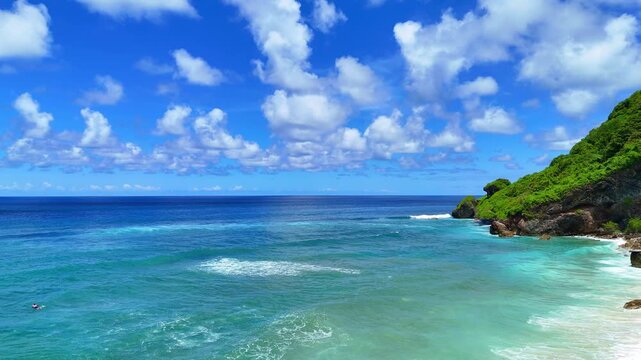 Breathtaking drone footage soaring over turquoise ocean waters along dramatic limestone cliffs in Uluwatu, Bali. Lush green hills and rugged coastline with crashing waves. Captured in 16:9 horizontal 