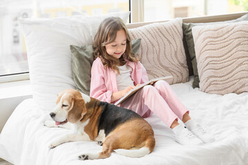 Obraz premium Cute little girl with Beagle dog reading book on bed in bedroom