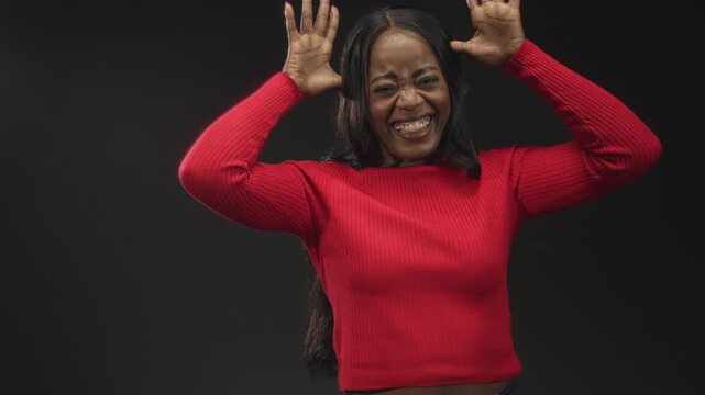 Woman in red sweater laughs, holding hands to head with fingers splayed, tongue out and eyes squinting in a dark studio portrait, playful expressive gesture; joy playfulness.