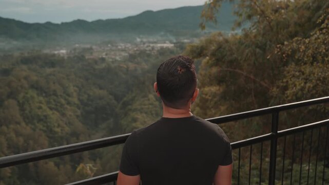 A man stands on a viewpoint overlooking misty forest mountains during early morning. Shot at Tebing Keraton, Bandung, West Java, Indonesia. Concept of solitude, reflection, and connection with nature 