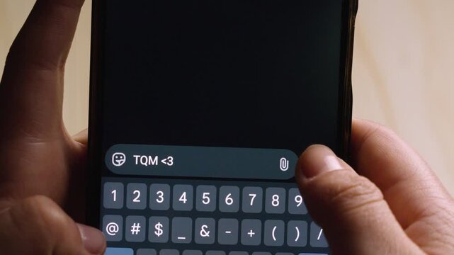 Close-up of hands typing the Spanish abbreviation "TQM" (Te Quiero Mucho) on a smartphone. 