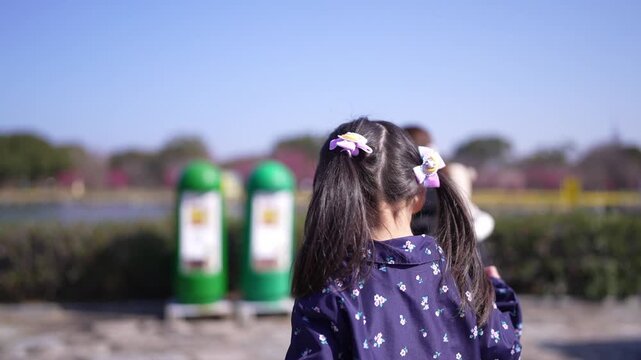 Little girl walks with mother near lake. Her hair adorned with small flowers. Child dressed in patterned jacket, curious, exploring. Parent walks beside, guiding, supporting