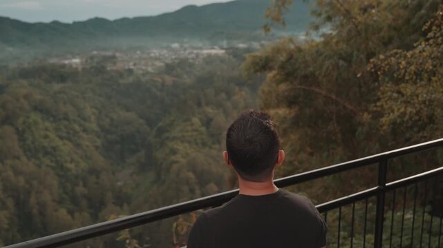 A man stands on a viewpoint overlooking misty forest mountains during early morning. Shot at Tebing Keraton, Bandung, West Java, Indonesia. Concept of solitude, reflection, and connection with nature 