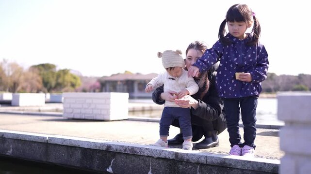 Bright day, three children on a park dock. One child points skyward, another sits close. Young girl smiles, holding baby in arms. Family moment captured near calm water