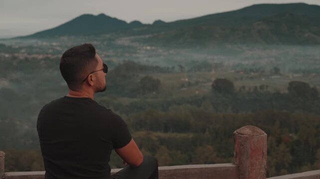 A man stands on a viewpoint overlooking misty forest mountains during early morning. Shot at Tebing Keraton, Bandung, West Java, Indonesia. Concept of solitude, reflection, and connection with nature 