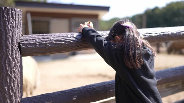 Little girl grips fence, eyes fixed on animals. She leans forward, curious, eager to see. Background holds soft light, wooden fence, dirt. Zoo or farm setting, gentle, natural atmosphere
