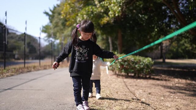 Little girl walks outside, holding a green ribbon. Her arms stretch as she moves with joy. Sunlight shines through autumn trees behind her. She smiles at her play, unbothered by distance