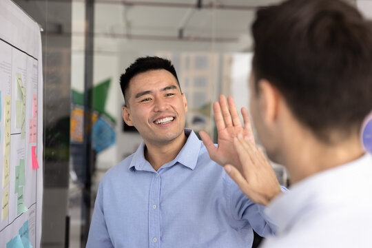 Agile teamwork success. Excited young businessman scrum master data analyst high five male coworker near planning board with colorful memo notes celebrate finishing sprint solving challenging problem