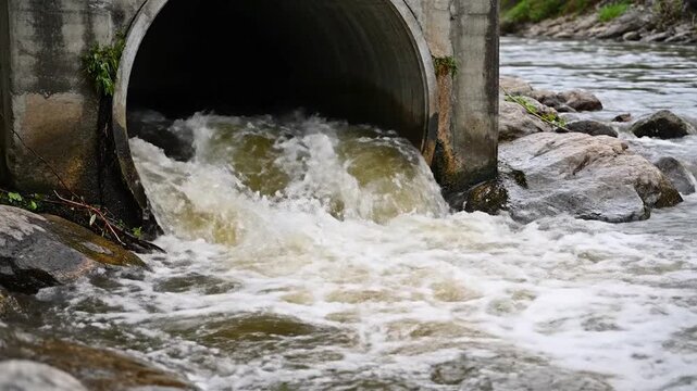 A large concrete pipe discharges murky water into a flowing river, creating turbulent rapids and white foam around surrounding rocks