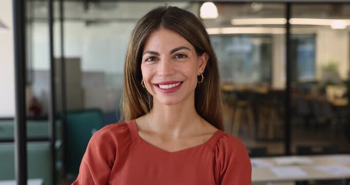 Positive beautiful 30s Latin businesswoman in casual posing for head shot, looking at camera, getting happy, smiling, standing in office. Hispanic company manager, business leader woman portrait