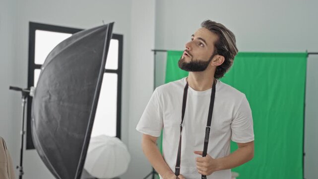 Man photographer holds camera strap and raises camera to eye in studio with green screen and softbox lighting visible; concentration creativity.