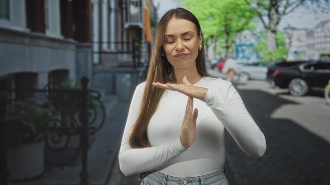 Young woman making timeout hand gesture with palms visible, wearing white top and jeans on a cobblestone street near parked cars; frustration restraint.