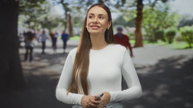 Woman holding vaporizer near mouth, index finger raised, standing on a park path with trees and blurred pedestrians; confidence casual leisure.