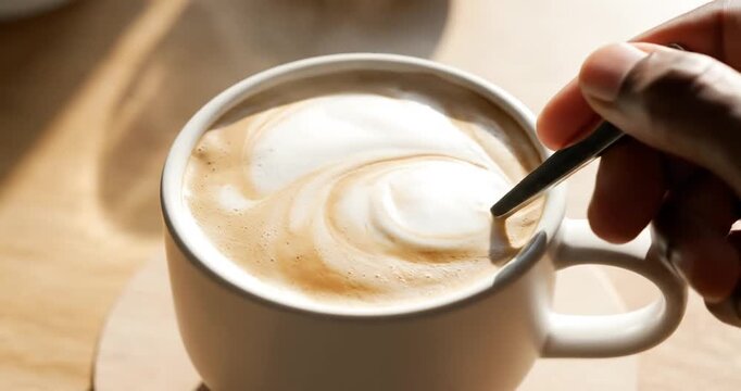 A close-up of a hand stirring a frothy coffee cup in soft morning light on a wooden table