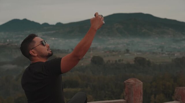 Traveler using smartphone to take photos and selfies with a misty mountain forest landscape. Filmed at Tebing Keraton, Bandung, West Java, Indonesia. Concept of modern travel, social media, and captur