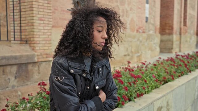Teen woman with curly hair stands with arms crossed beside a stone building and red flowerbed; melancholy solitude.