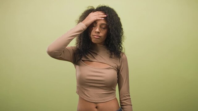 Young woman with curly hair touching forehead with hand, exposed midriff visible, wearing crop top in studio with neutral green backdrop; frustration fatigue.
