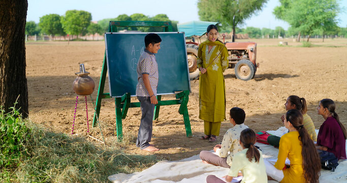 Indian happy young adult gen z female do work talk to villager girl boy sitting on mat rural area. teen male child writing study look board enjoy desi life open day class field ground place