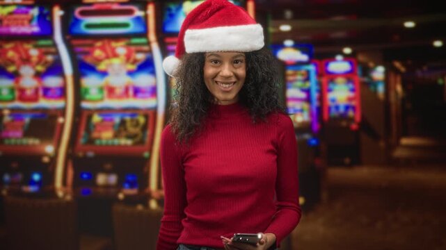 Woman with curly hair wearing a santa hat touches the hat brim with her right hand while holding a smartphone, slot machines visible behind her in a building; festive smile.