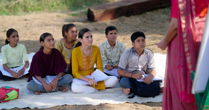 Indian mature adult gen z female lady talk speak share good news teach to villager group day under tree. Happy girl boy sitting on mat do study look board learn at open class field rural area place