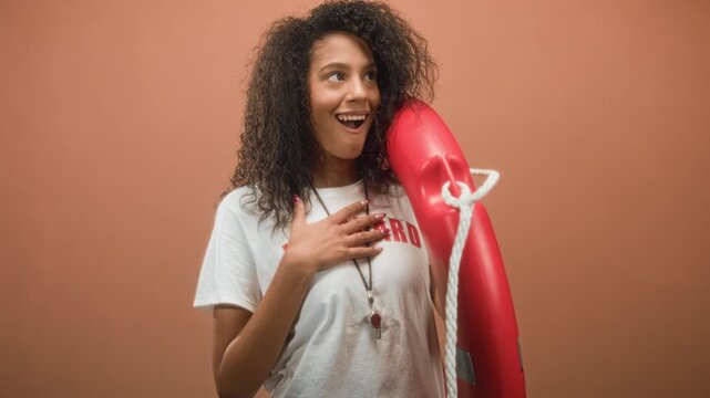 Woman lifeguard holding red rescue buoy with whistle on lanyard, hand on chest smiling in studio; pride duty.