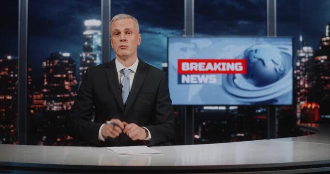 Professional Man Journalist Sits at Desk, Gesturing With Pen While Looking at Camera and Speaking in Studio With Large Screen and Panoramic Night City Background. Concept of Evening News Broadcast.