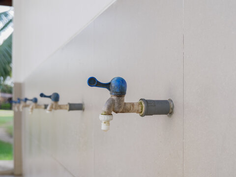 ​Row of outdoor metal water taps with blue lever handles mounted on a tiled wall. Public washing or ablution facility with a shallow depth of field