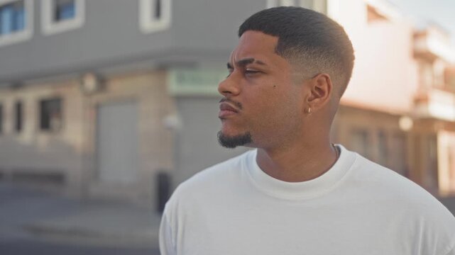 Man with beard and goatee looking left in street wearing white tshirt and earring, short fade haircut near storefront building; quiet contemplation.