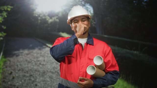 Construction worker man in hard hat holding rolled blueprints and covering mouth with hand on forest path; determination duty.