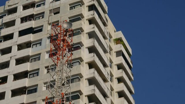 Tall white concrete residential high-rise building beside an orange lattice telecom transmission tower against a vivid blue sky