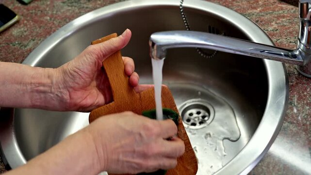 Hands Washing Wooden Cutting Board Under Running Water in a Kitchen Sink