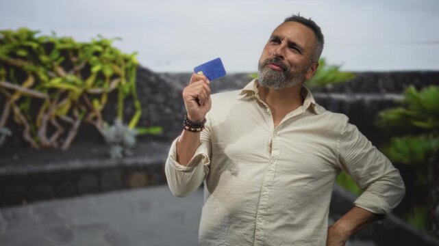 Man holding creditcard with finger on chin and bracelets visible, wearing beige shirt in street; financial decision thoughtful.