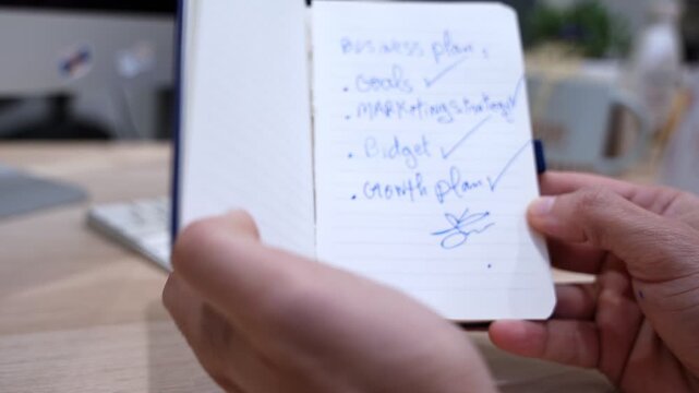Close-up of hands holding an open notebook with handwritten business plan notes, including goals, marketing strategy, budget, and growth plan in a modern office setting.