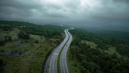 Fototapeta premium A dramatic aerial view of a highway in India winding through lush green hills and dense forest under a cloudy sky. Vehicles travel along the smooth, curving road, highlighting modern infrastructure.