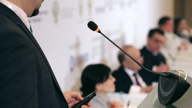 Business speaker talking into microphone and adjusting it during conference presentation in indoor event setting with audience in background.