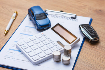 Obraz premium Car insurance document on clipboard with white calculator, stacked coins, blue toy car and modern car key on wooden desk. Concept auto insurance premium calculation and vehicle finance planning.
