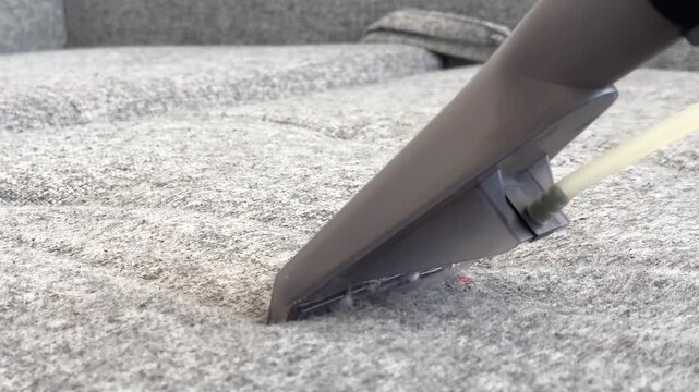 The person uses a vacuum attachment to clean stains from a grey couch