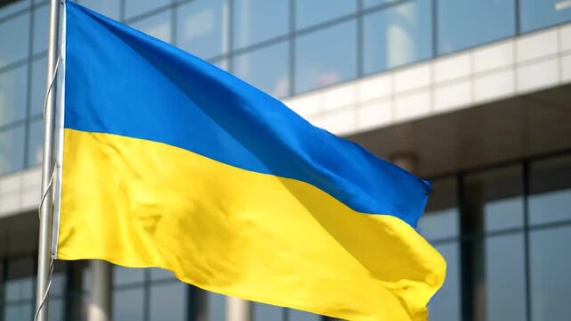 A blue and yellow flag waves gently in the breeze in front of a modern glass building