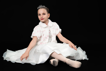 Elegant little girl in white ballet costume and tiara sitting gracefully on floor. Studio portrait of ballerina on black background with copy space. Represents childhood dreams, dance and performance © Alexander Piragis