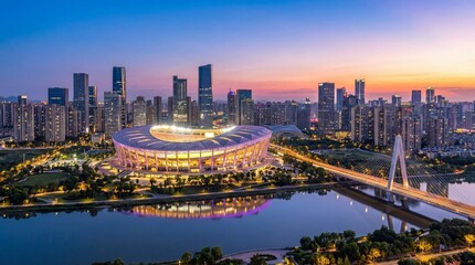 Fototapeta premium Panoramic view of a modern city skyline with a brightly lit stadium, skyscrapers, and a bridge over a river at sunset.